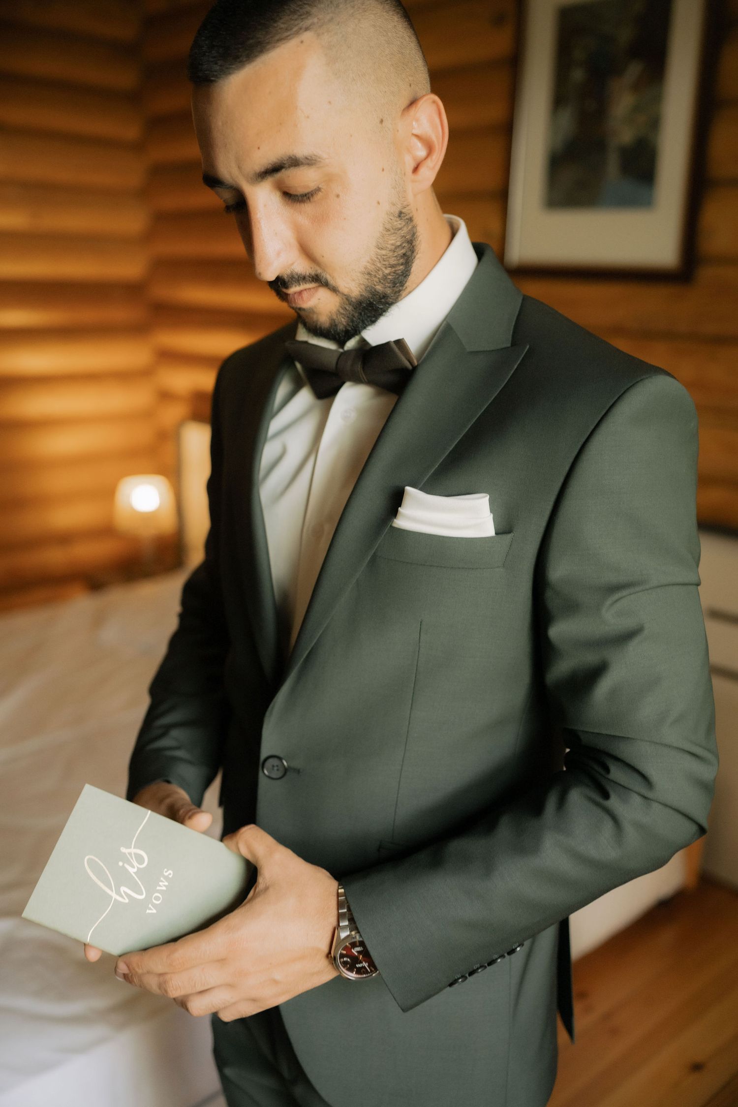 Groom in dark suit reads emotional letter before wedding ceremony in rustic wooden room.