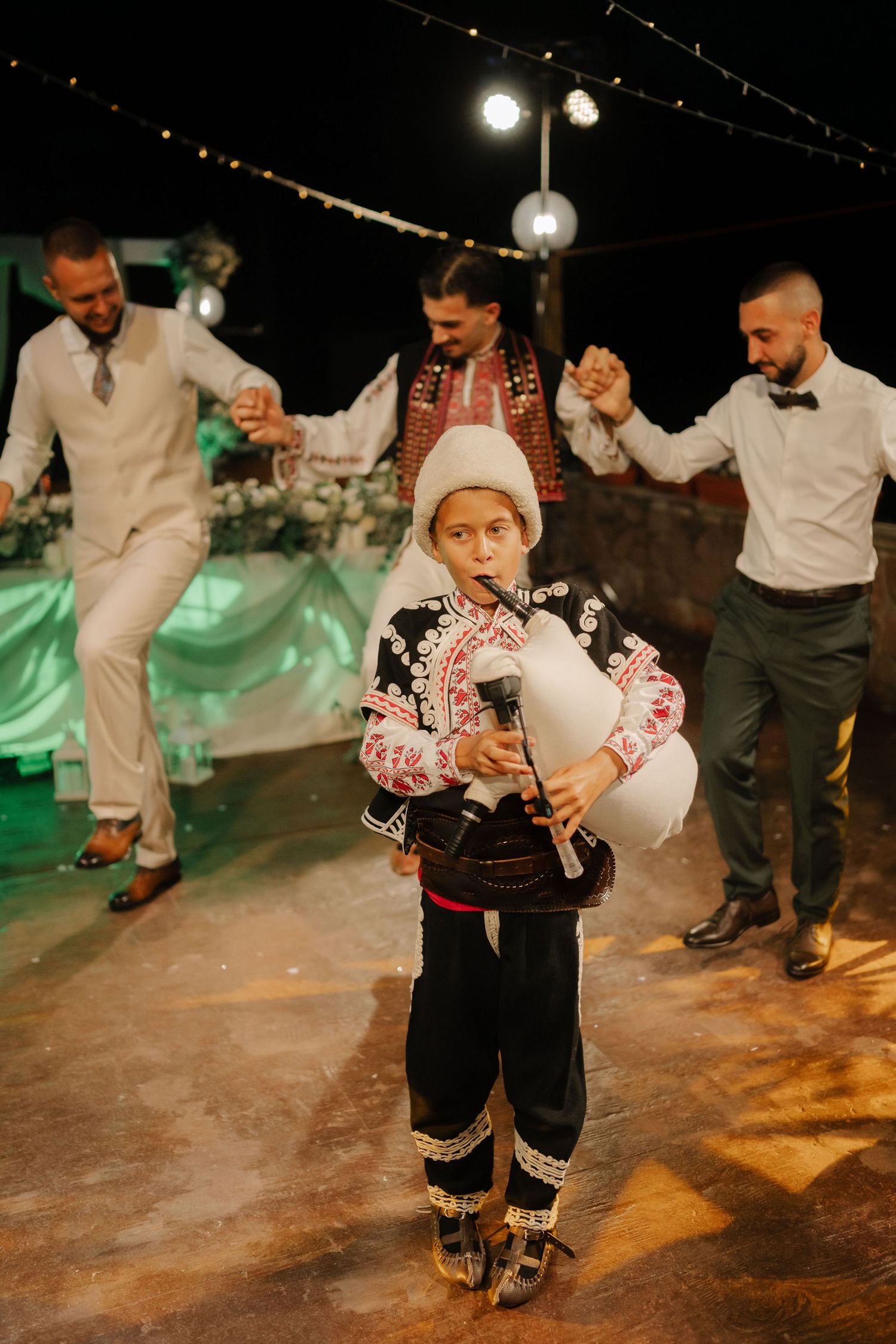 Traditional folk dancers in white shirts and dark pants perform energetic dance moves at a nighttime celebration.