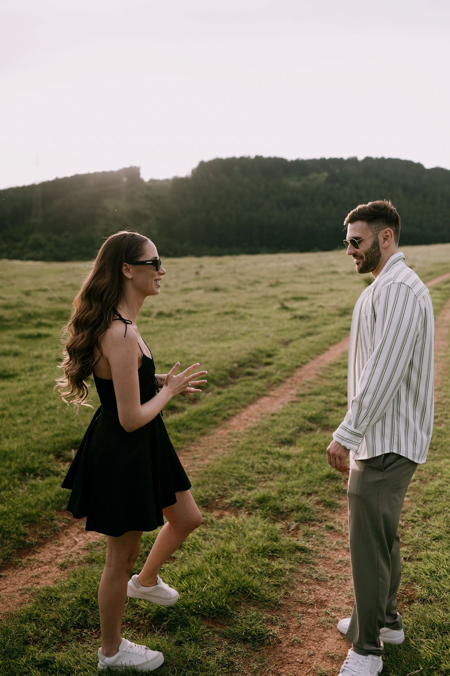 A couple walks along a grassy hillside path at sunset, wearing casual summer outfits and enjoying the golden hour.