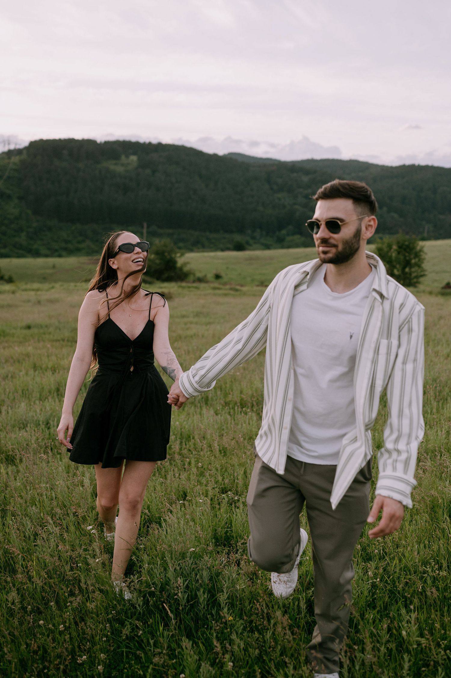 A couple playfully walks through a wildflower meadow with mountains in the background on a cloudy day.