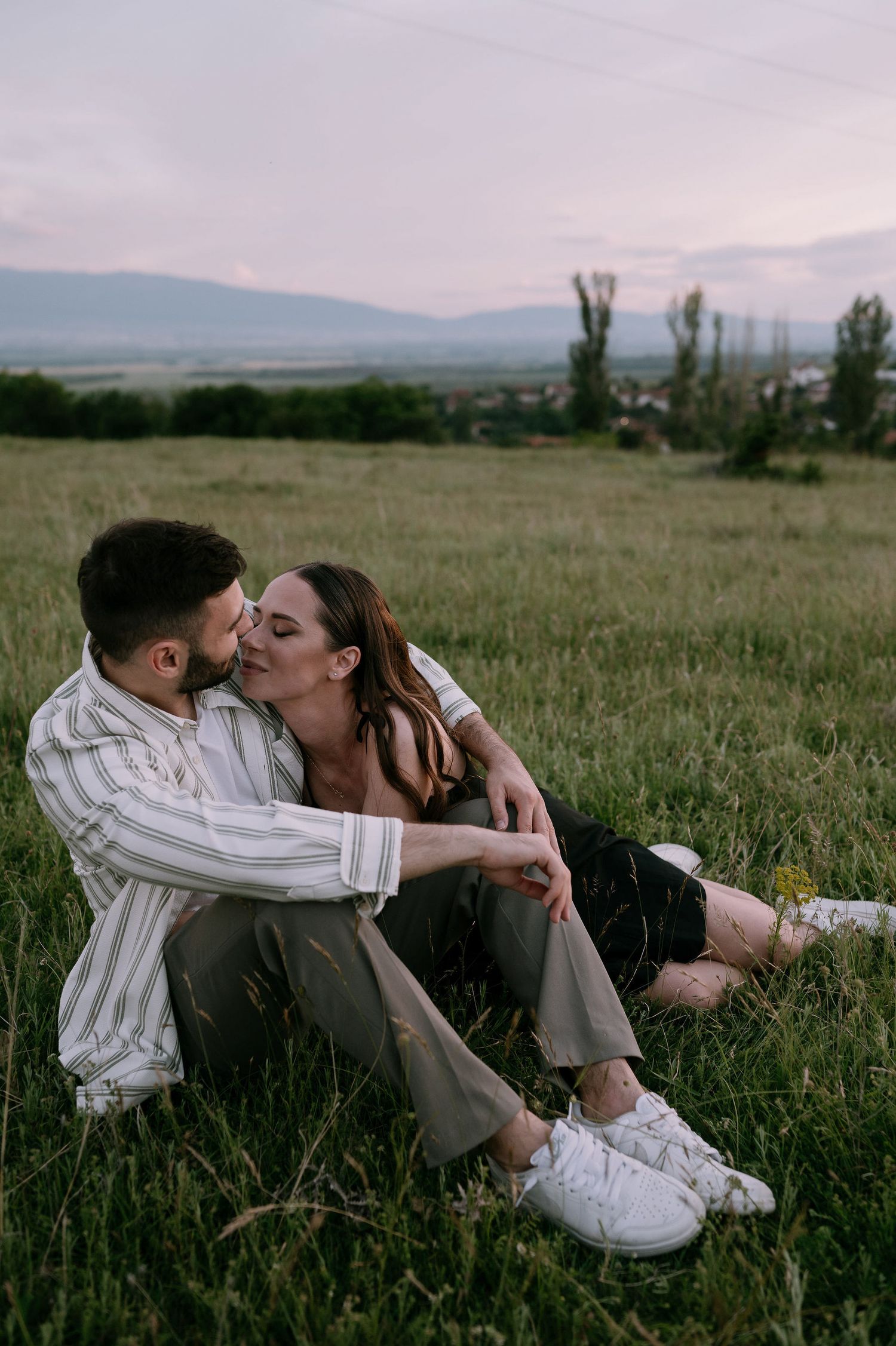 Romantic couple sharing intimate moments while lying in a grassy meadow with mountains in the background.