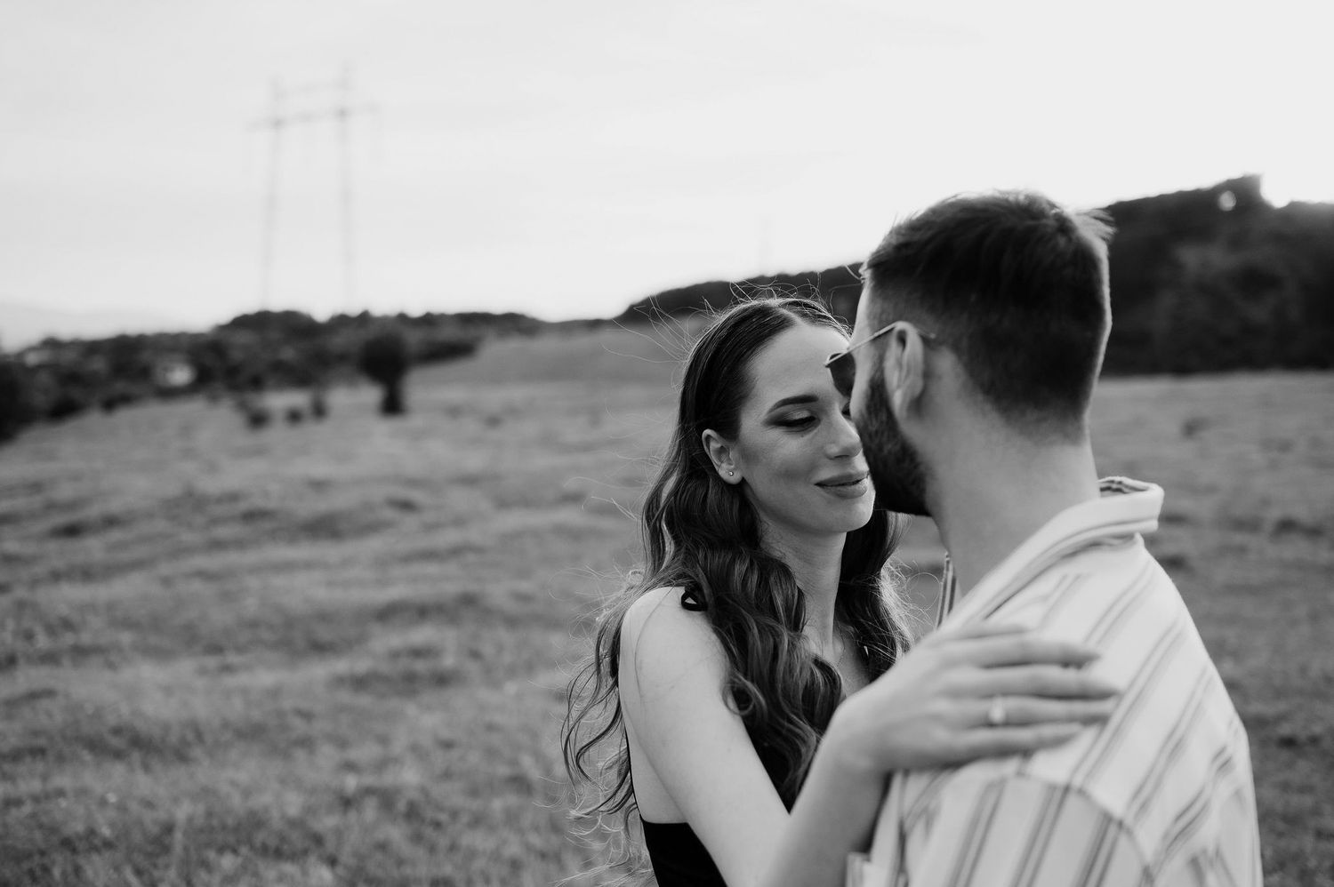 Black and white romantic moment between couple embracing in an open field with power lines in background.