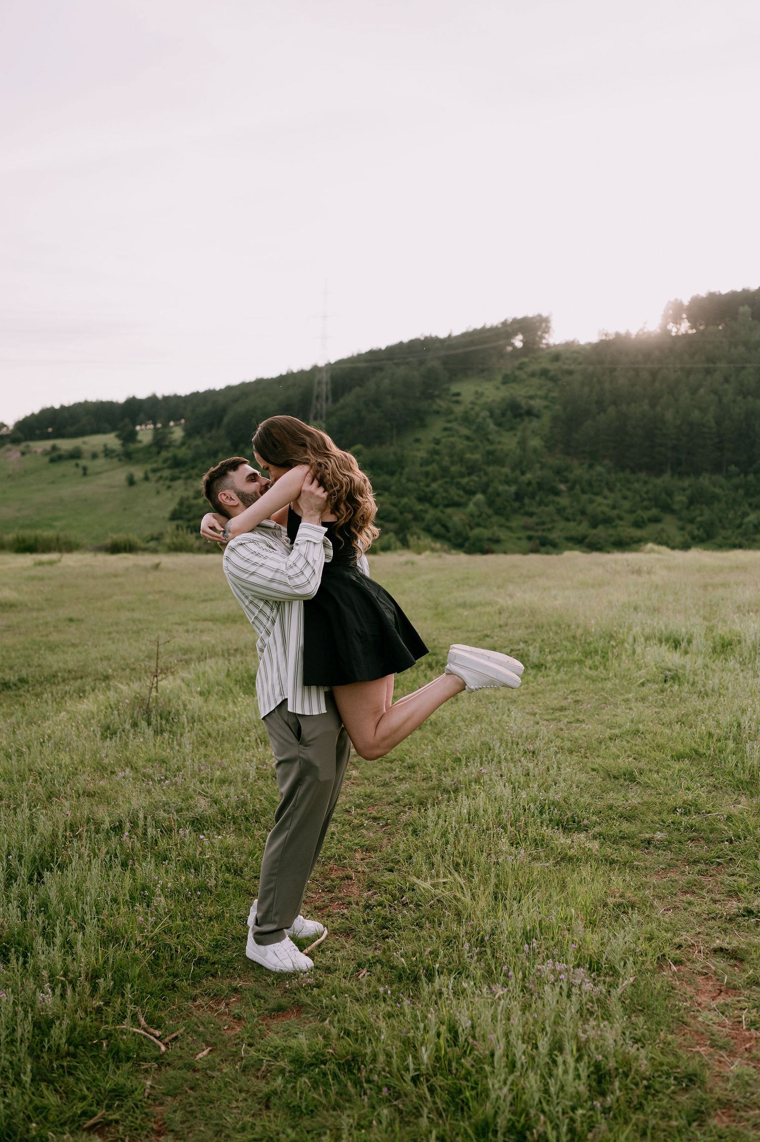 A couple embraces and spins playfully in a lush green meadow at sunset.