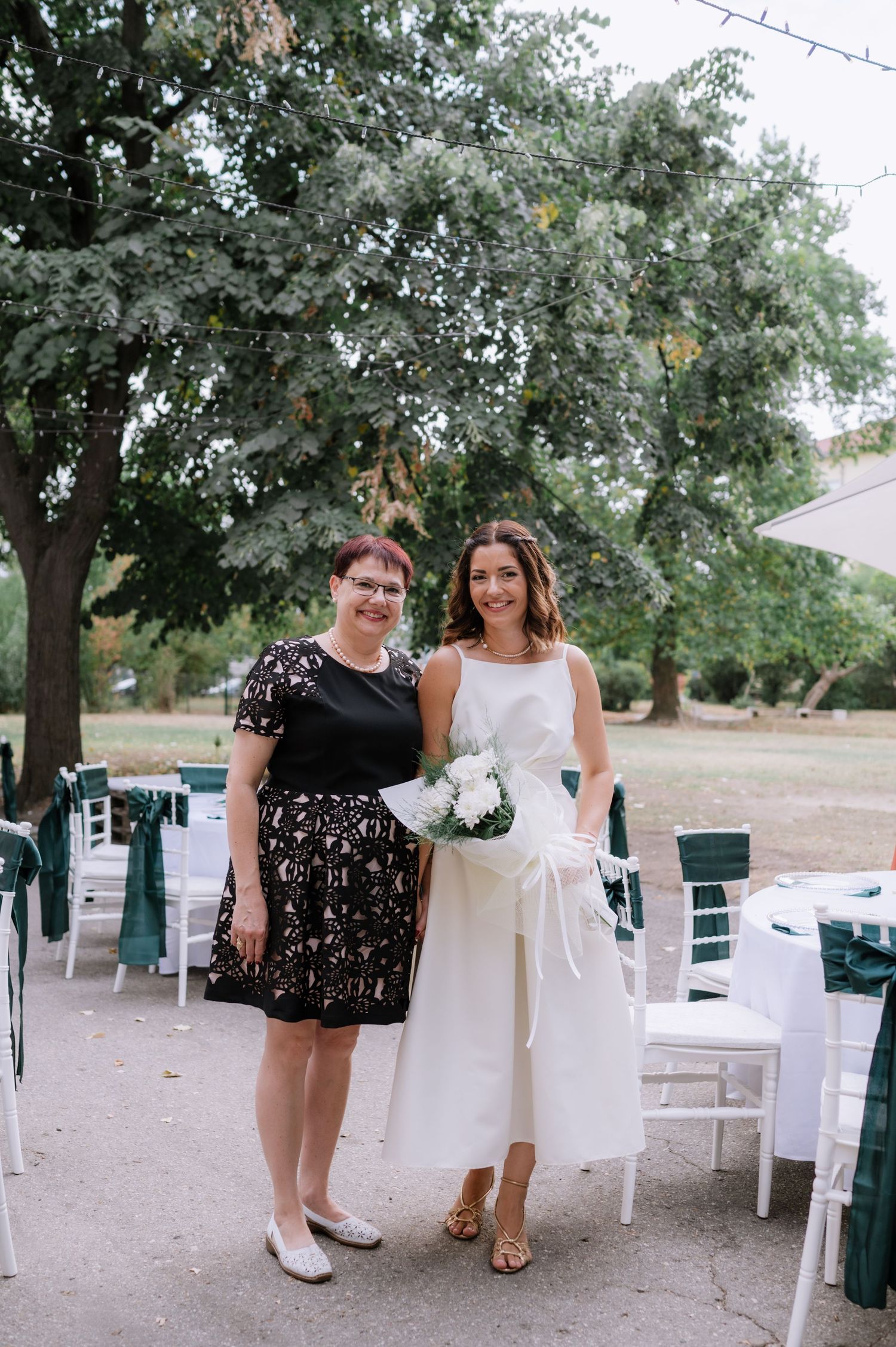 Two people in wedding attire sit on white chairs outdoors under shady trees at a garden ceremony.