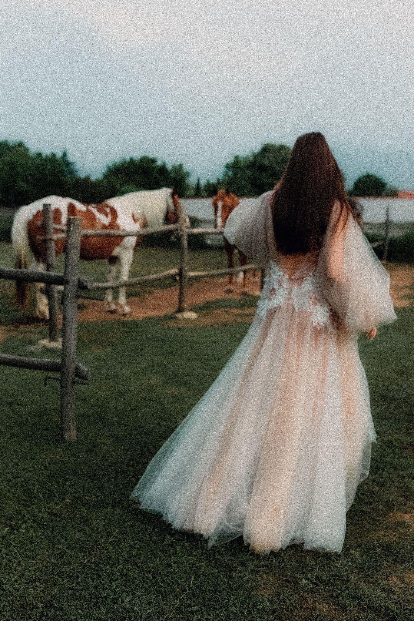 A bride in a flowing white tulle gown poses gracefully near rustic wooden fences and horses in a pastoral countryside setting.