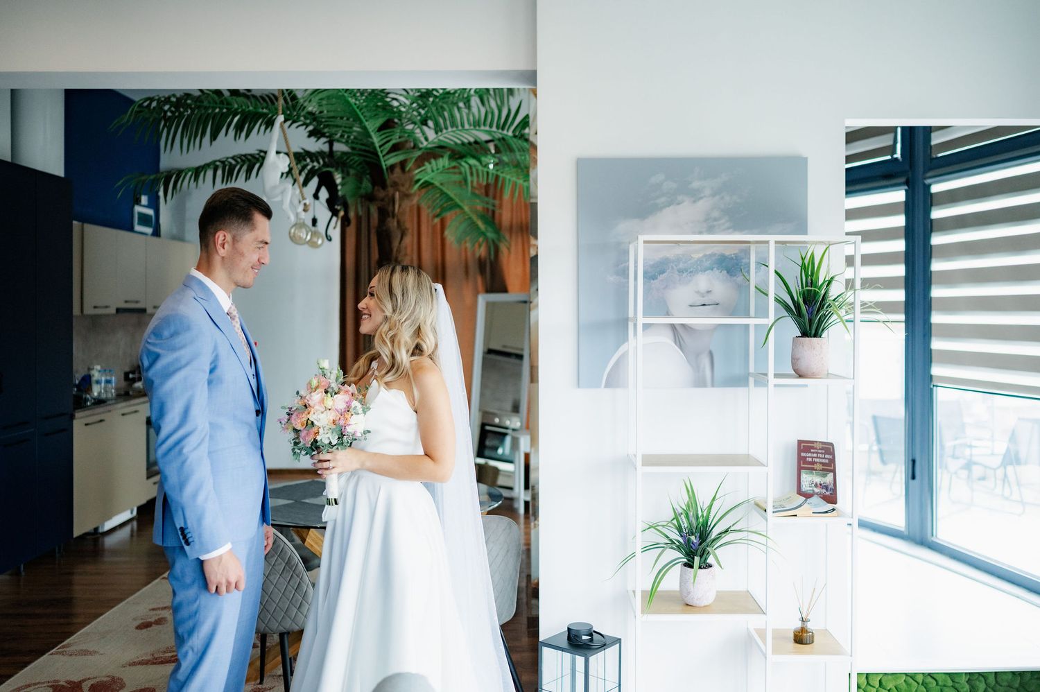 A couple in blue and white attire share intimate moments in a modern, bright interior space with plants and windows.