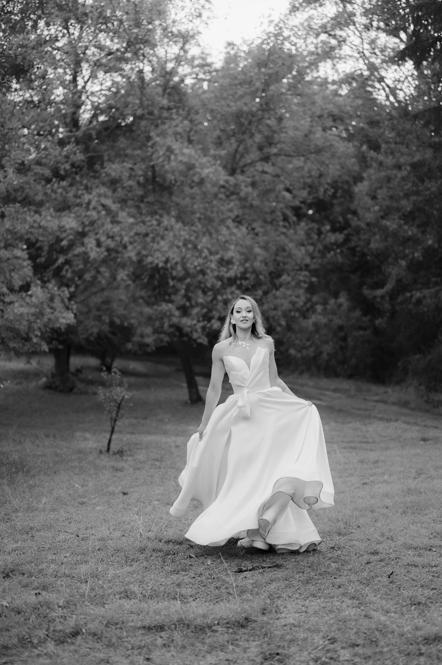 A graceful figure in a flowing white dress twirls in a meadow surrounded by trees in black and white photography.