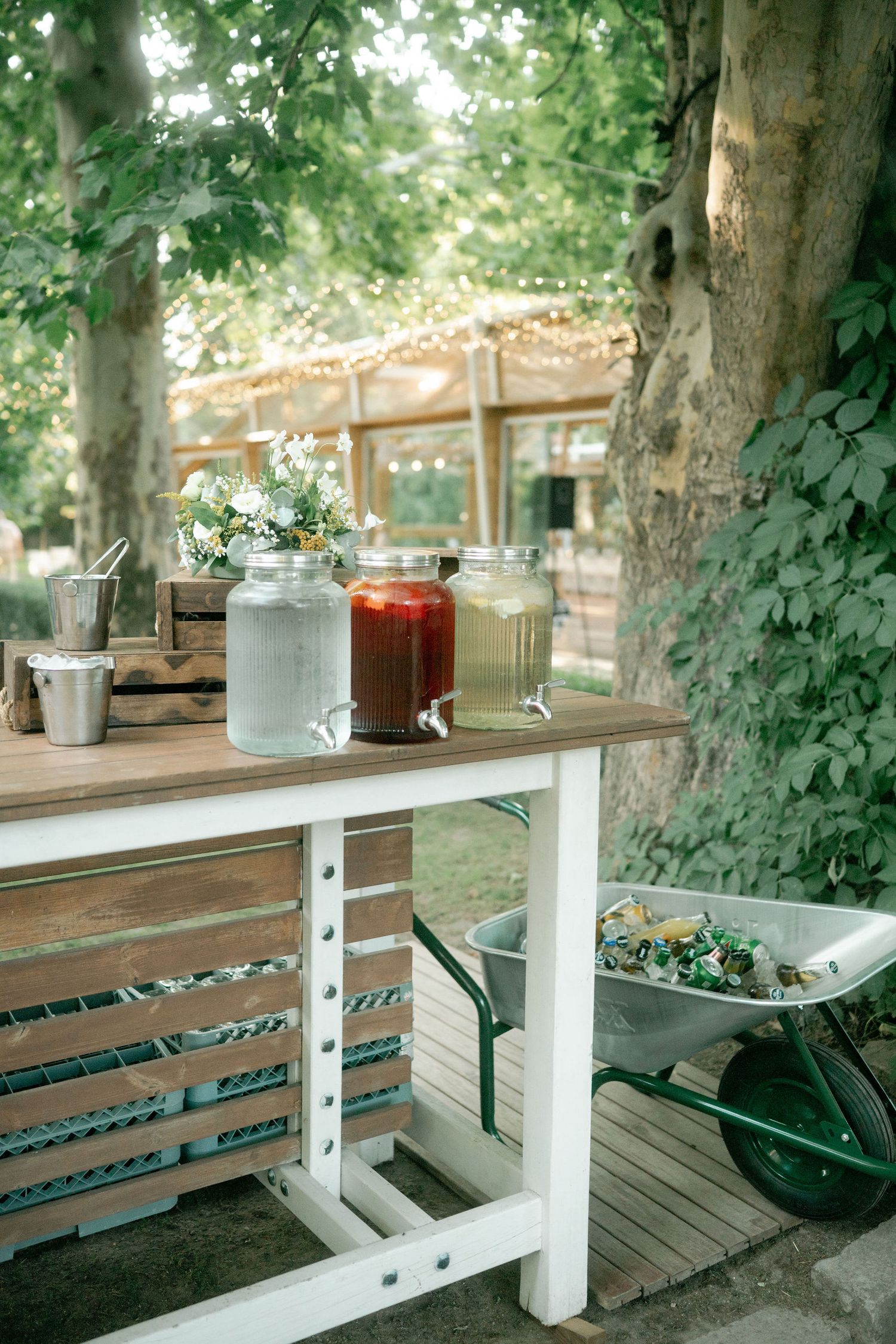 Rustic outdoor seating area with white wooden benches and glass jars on a deck surrounded by lush green trees.