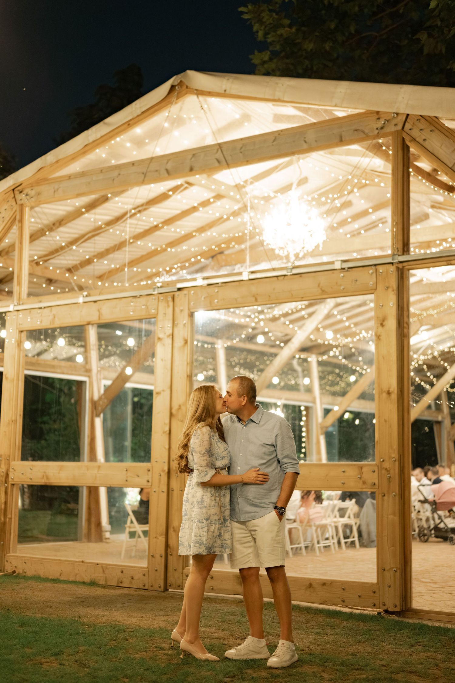 Clear wedding tent decorated with string lights and couples dancing on grass at dusk during outdoor reception.