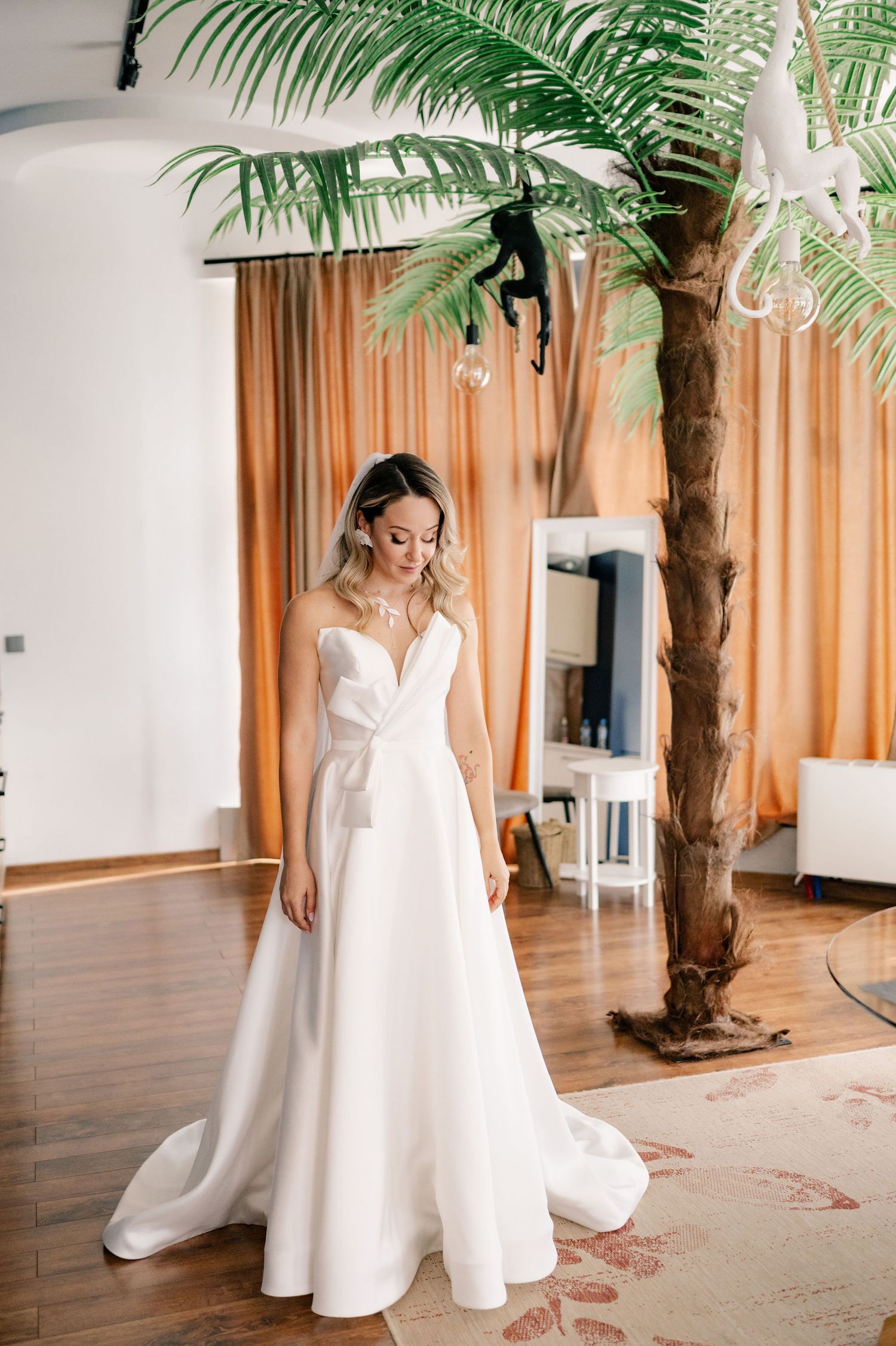 A flowing white wedding dress is displayed in a sunlit studio space with a decorative palm tree and orange curtains.
