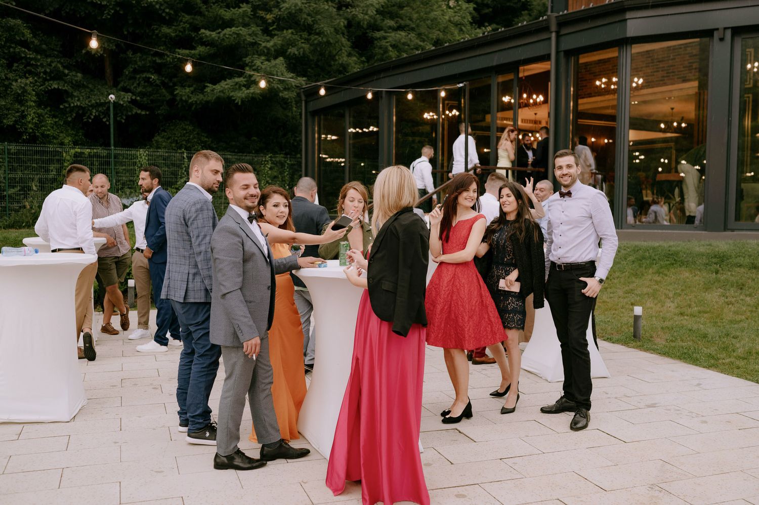 Wedding guests mingling and dancing outdoors on a stone patio surrounded by lush greenery.