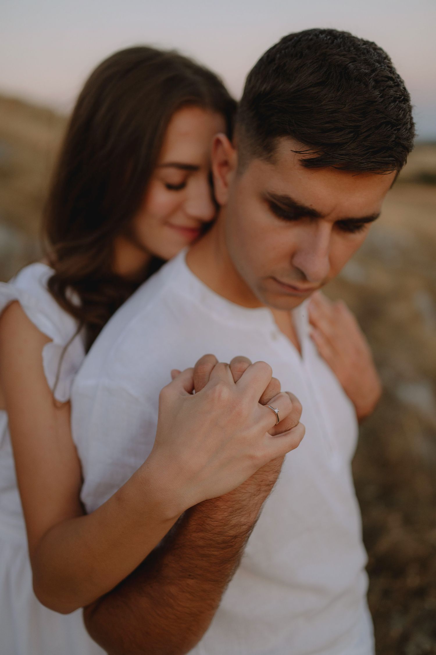 Intimate couple in white shirts embrace tenderly during golden hour photo session in natural outdoor setting.