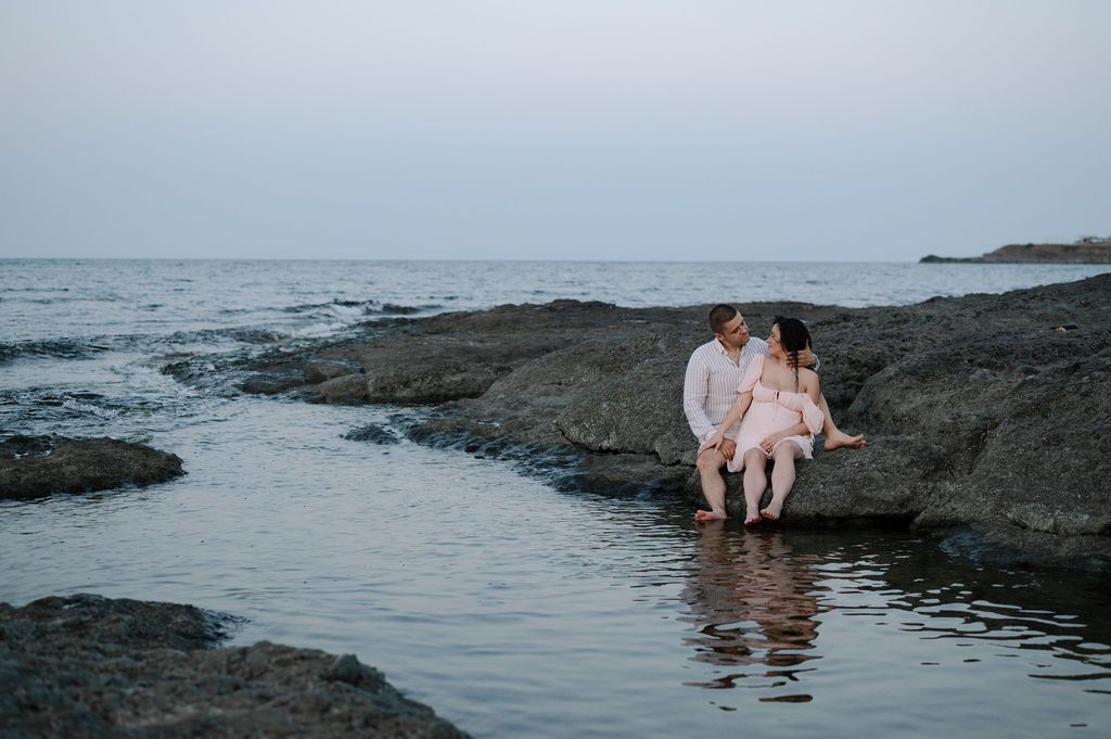 A couple sits together on rocky coastal cliffs at sunset overlooking the ocean waves.