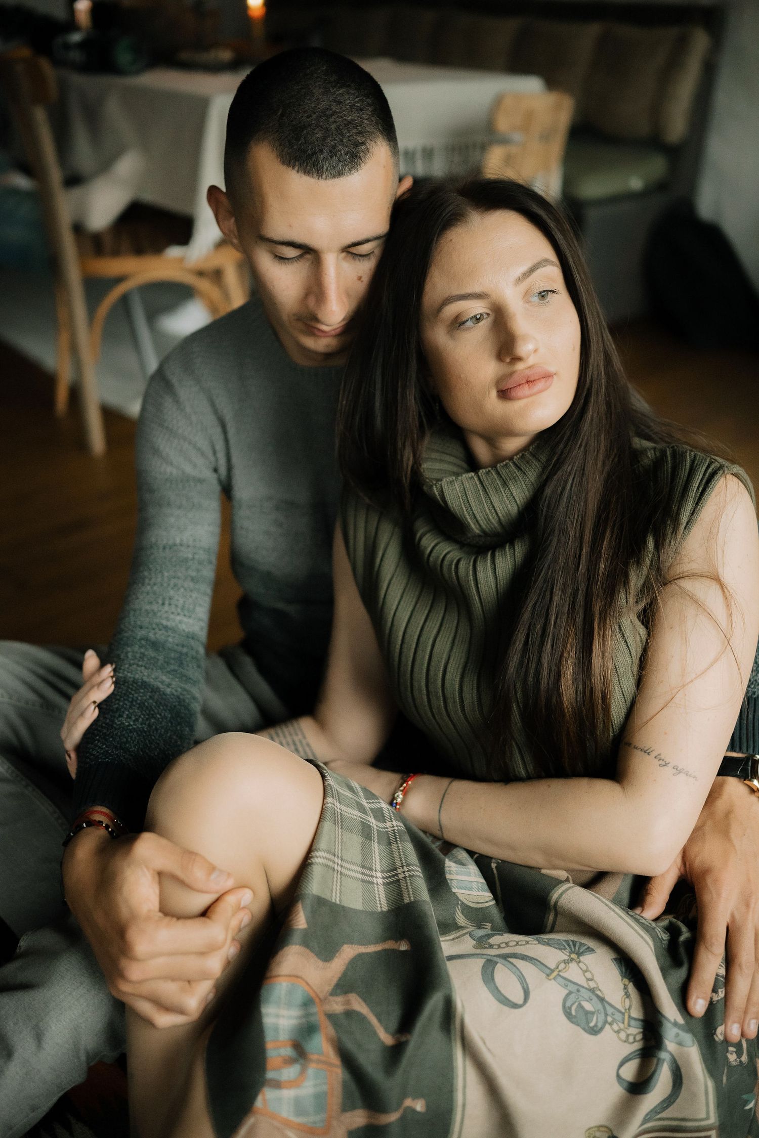 A couple sits close together on a couch in moody lighting, showing intimate body language and connection.