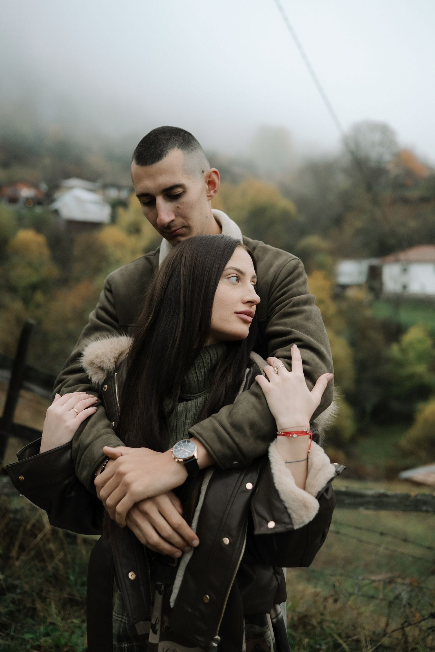 A couple embraces tenderly in an outdoor autumn setting with trees and mist in the background.