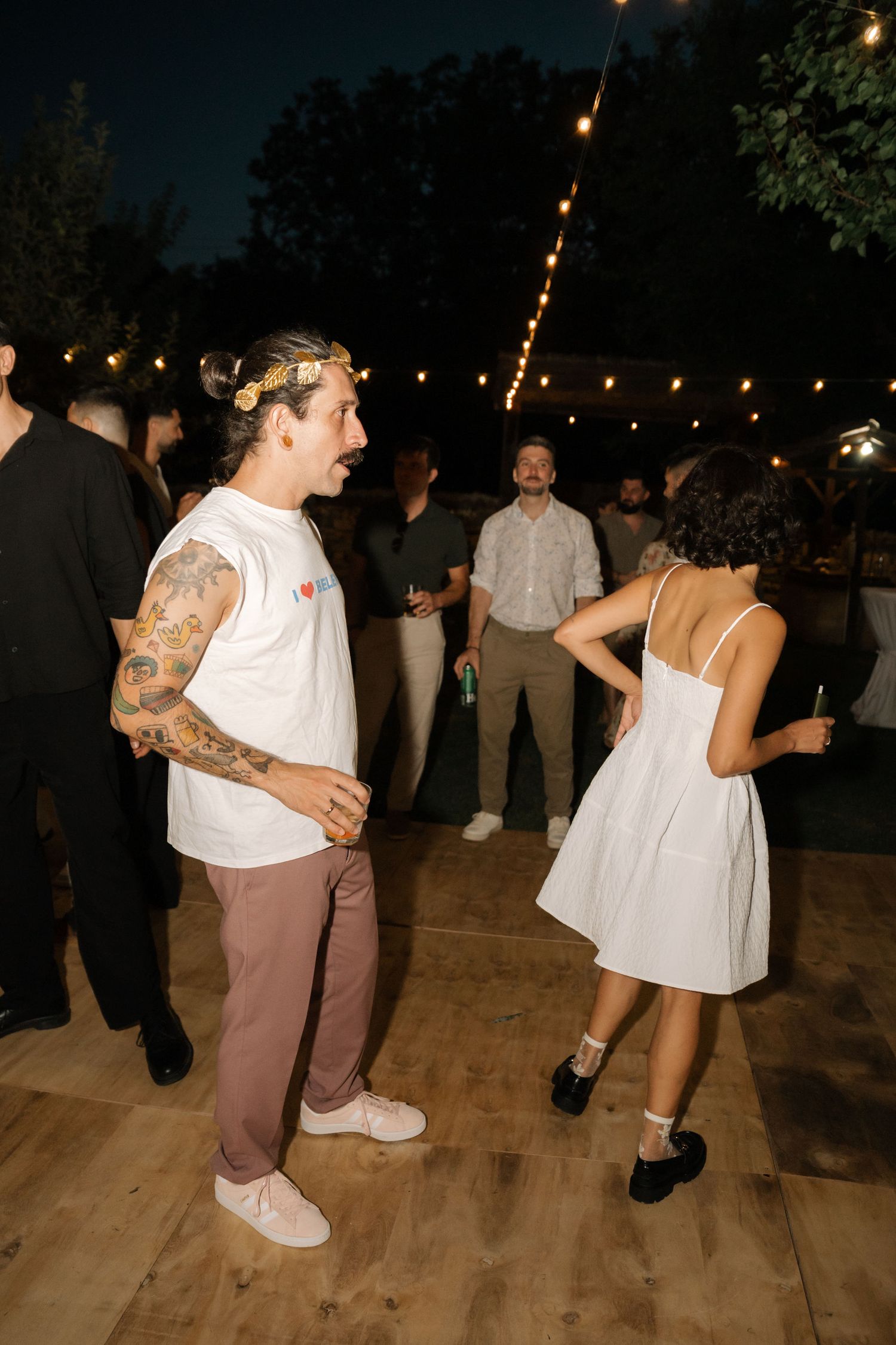 People dancing together on a wooden dance floor at a nighttime outdoor party with string lights overhead.