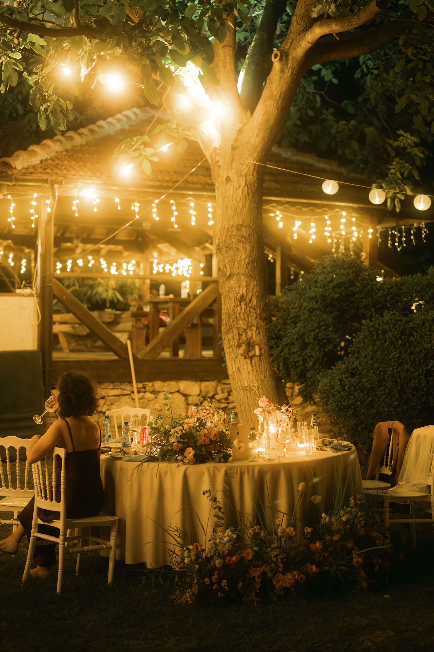String lights illuminate an outdoor evening wedding reception with round tables and chairs set beneath trees.