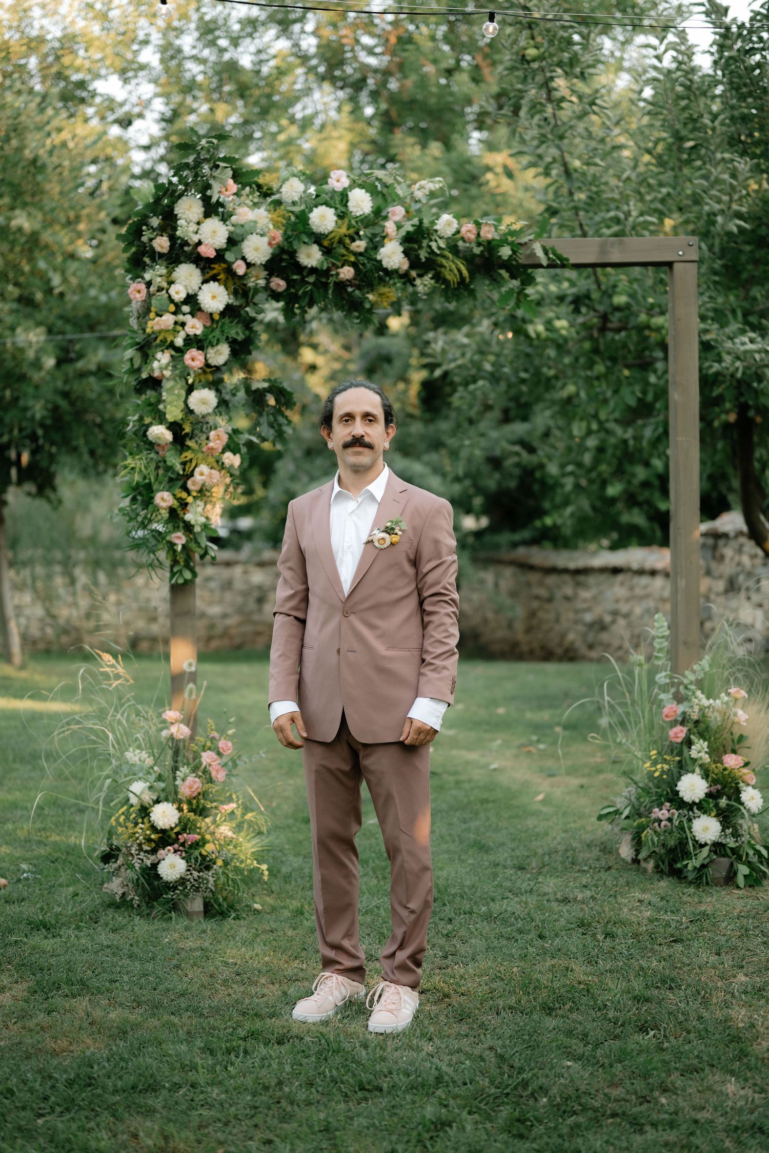 Wedding portraits in a garden setting with white roses and greenery featuring a mauve-suited figure.