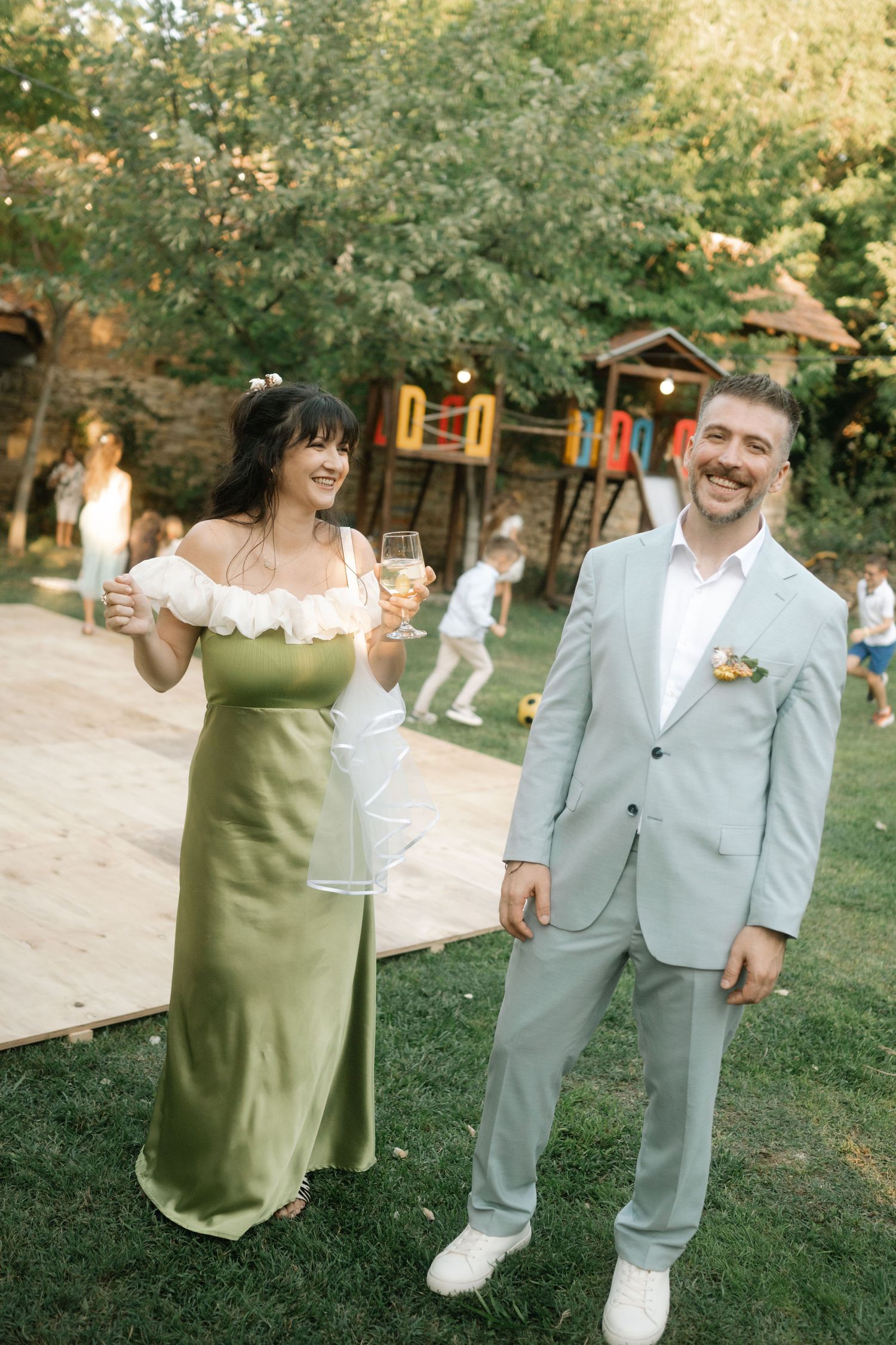 A couple in wedding attire poses playfully on a grassy lawn during an outdoor celebration.
