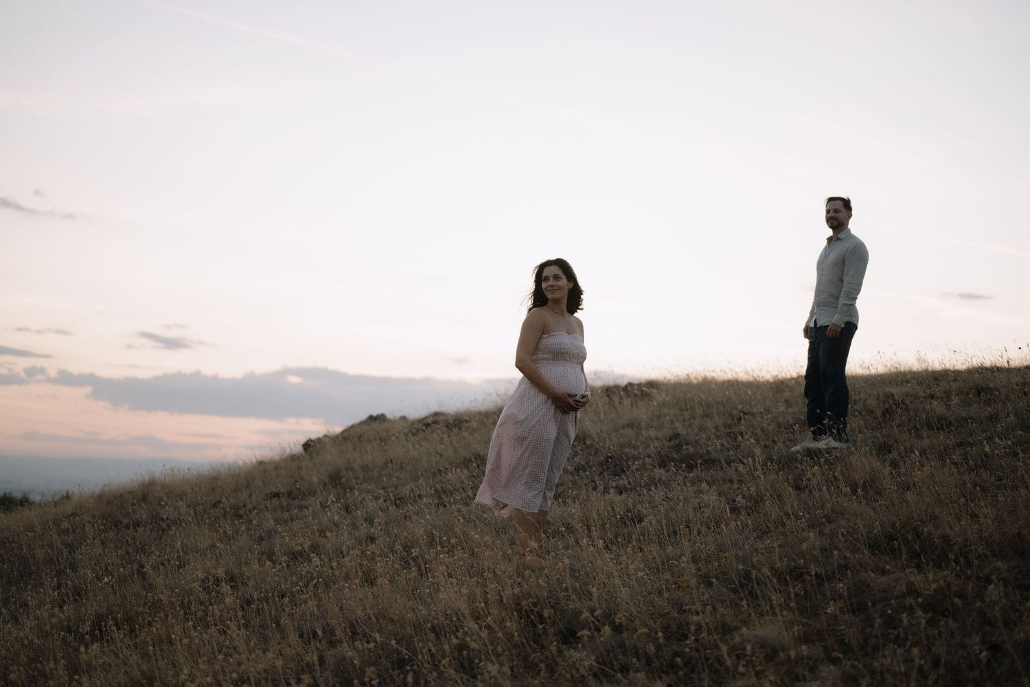Two people stand on a grassy hilltop at sunset, with one wearing a flowing white dress.