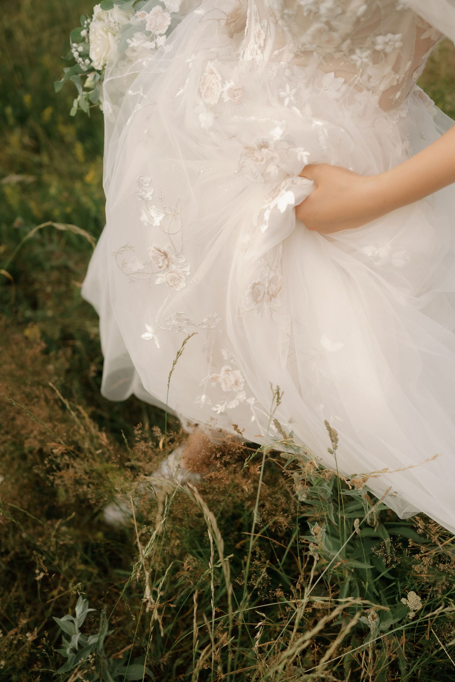 A flowing white wedding dress with delicate lace details rests gracefully among wild grass and foliage in a natural setting.
