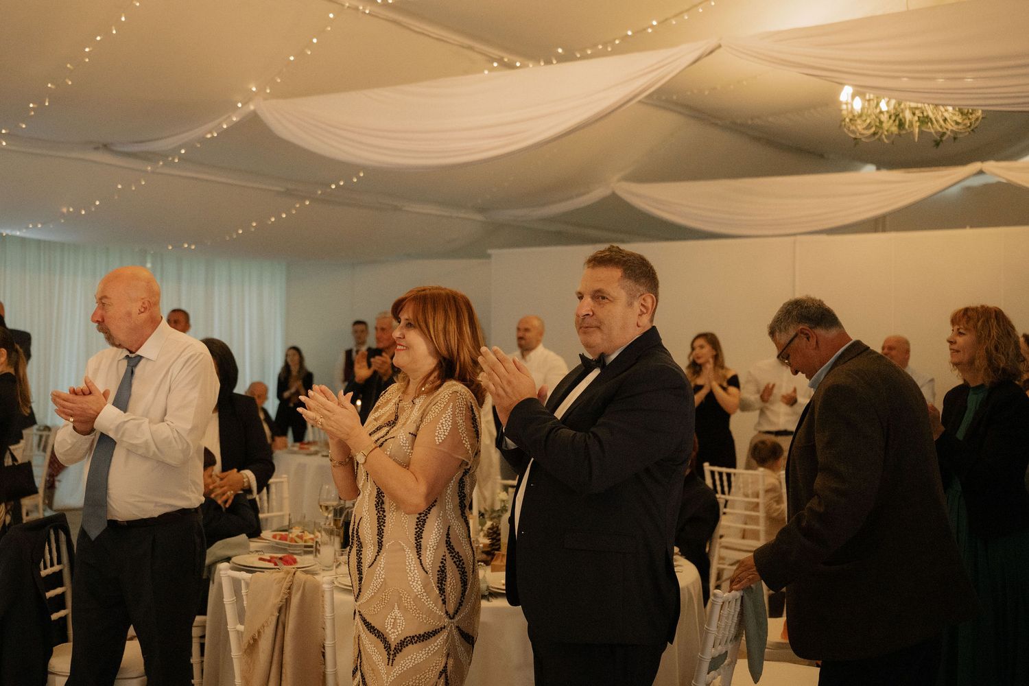 Guests applauding at an elegant wedding reception in a decorated venue with draped ceiling and fairy lights.