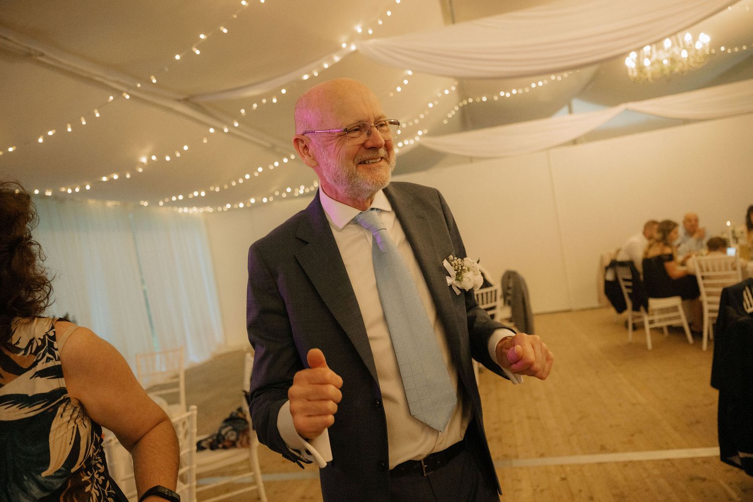 Elderly man in navy suit dancing at elegant wedding reception with fairy lights and white draping.