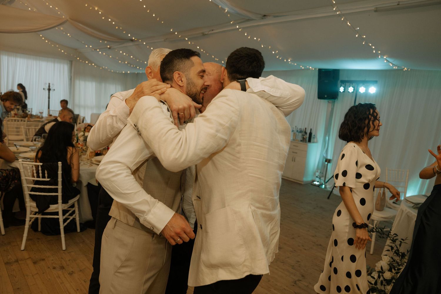 Two grooms share an emotional hug at their wedding reception, surrounded by guests in a decorated venue.