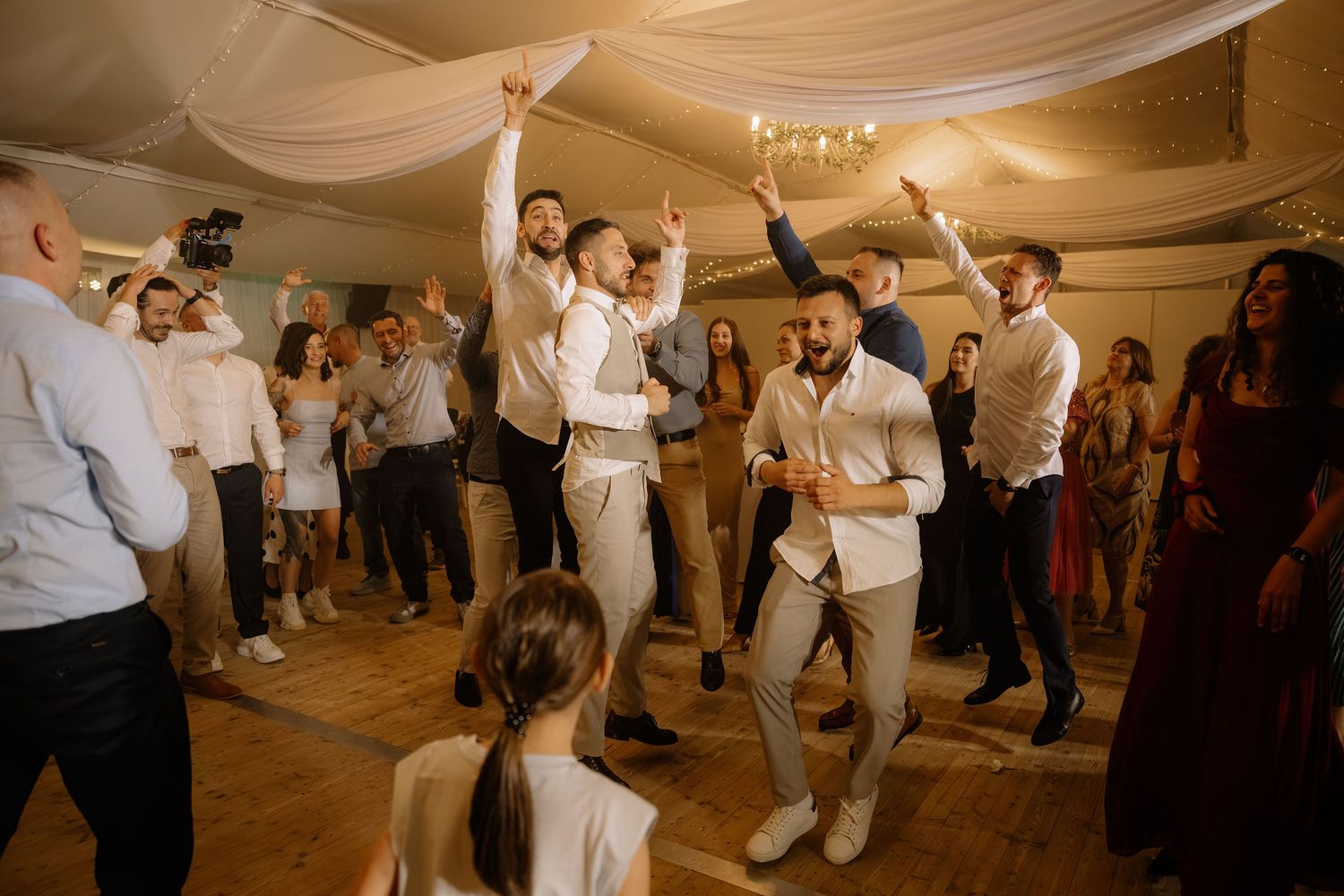 Guests dancing energetically at a wedding reception in an elegant venue with draped white ceiling decor.