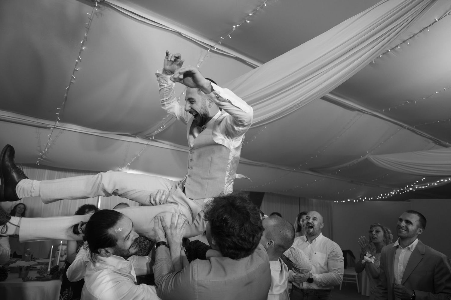 Black and white photo of groom being lifted by guests during lively wedding reception celebration.