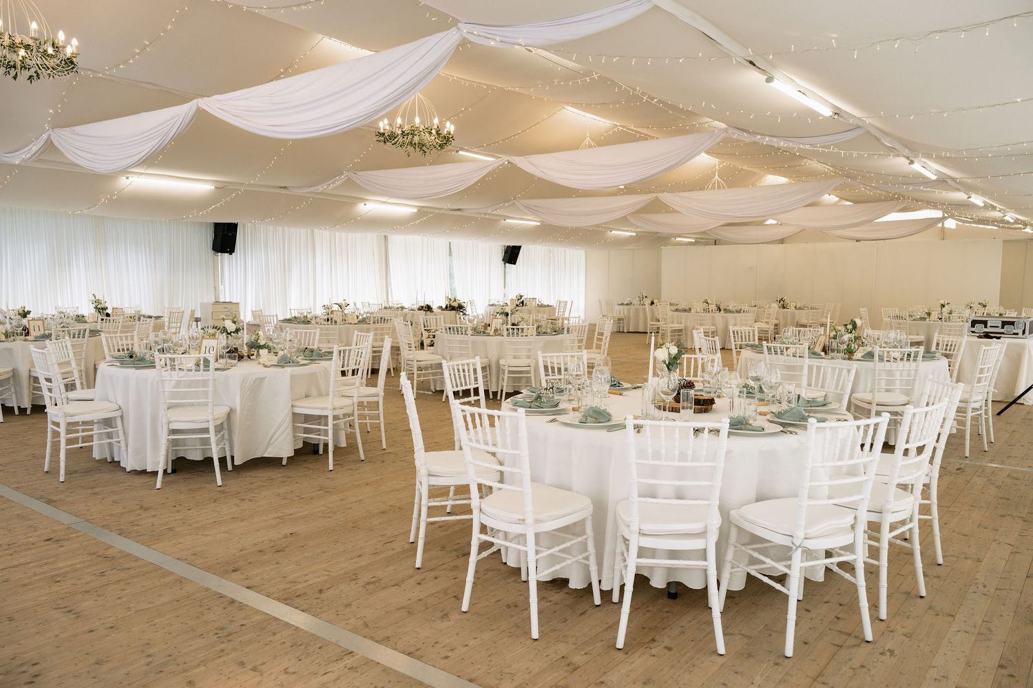 Elegant white wedding reception venue with draped ceiling, chandelier, round tables, and white Chiavari chairs on hardwood floors.