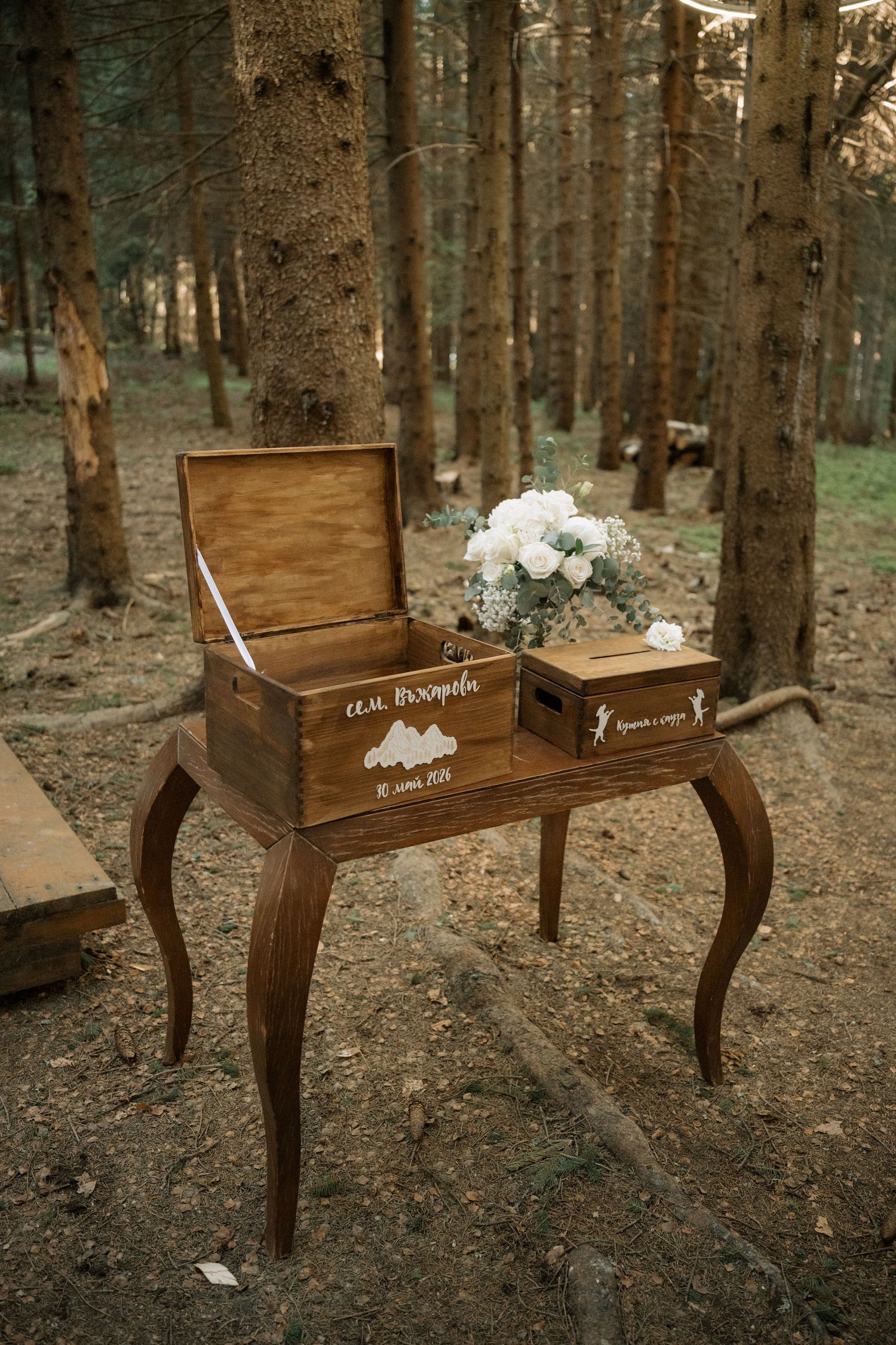 Rustic wooden wedding card box with white floral bouquet on elegant table in forest setting.