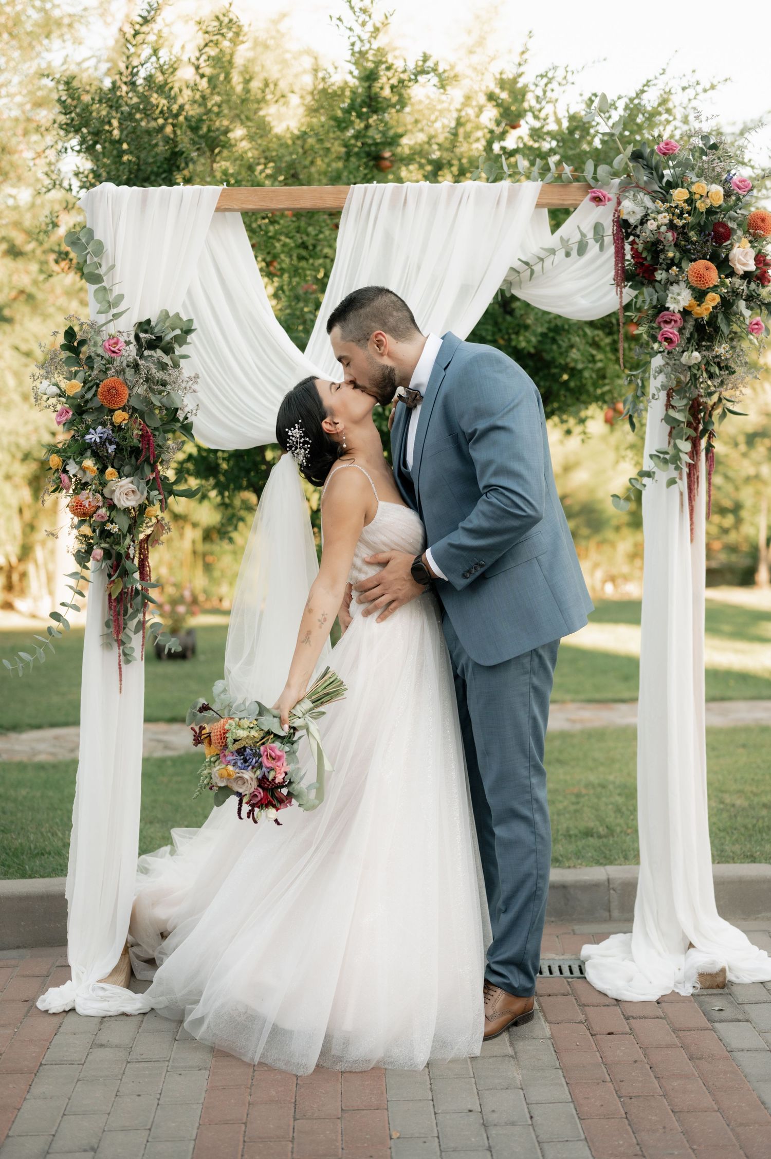 Romantic wedding couple sharing a kiss under a white draped arch decorated with vibrant floral arrangements.