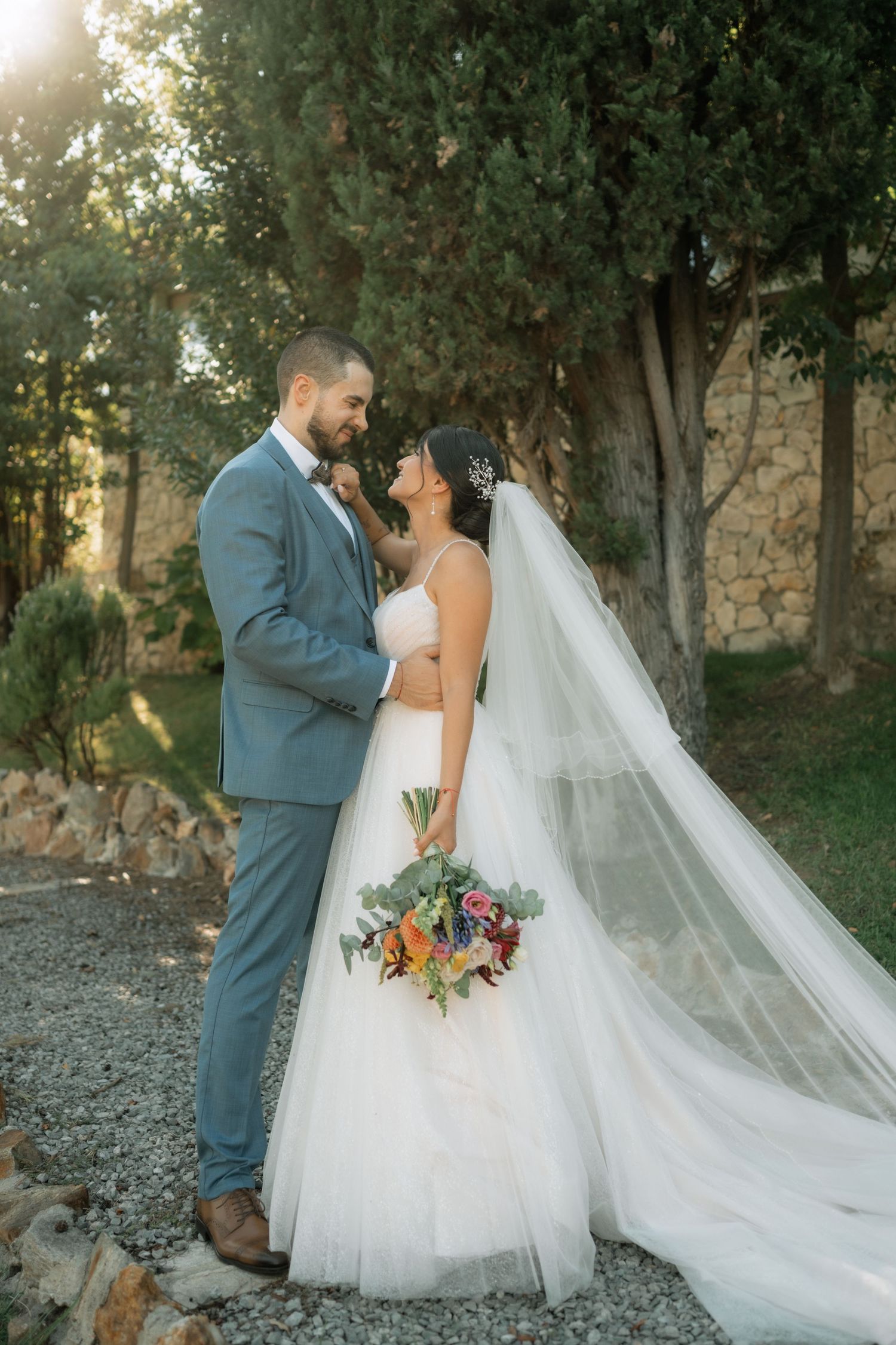 A romantic wedding scene in a wooded outdoor setting with a flowing white veil and gray suit at sunset.