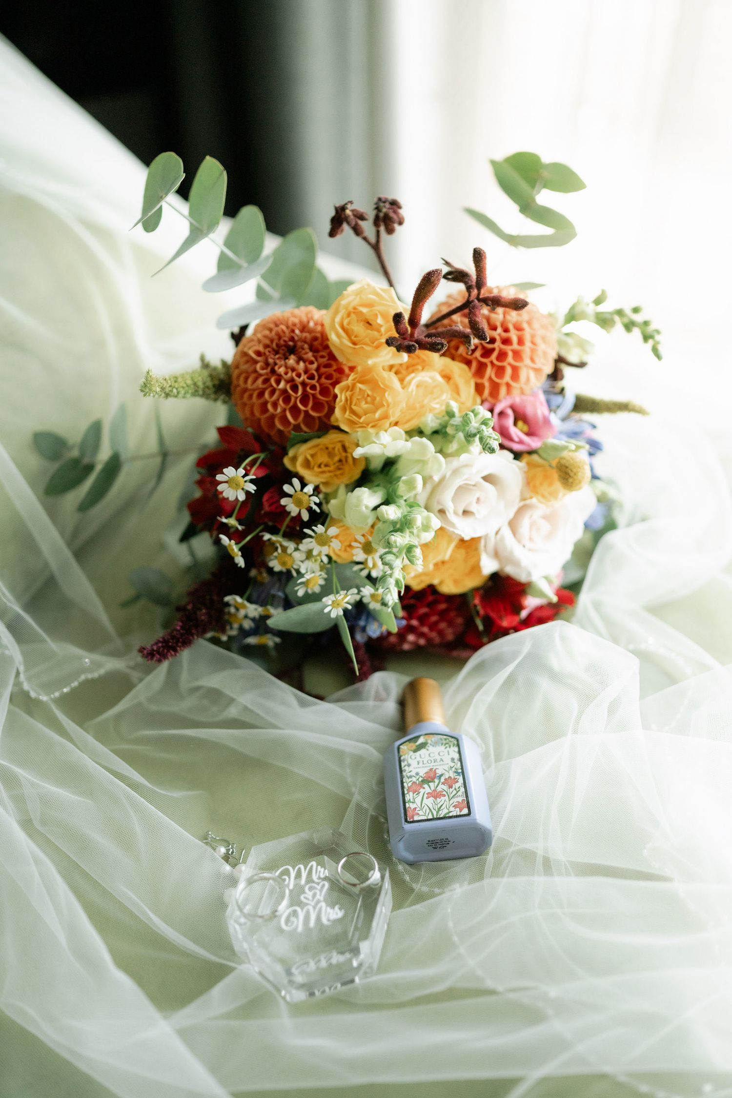 Elegant floral wedding details with orange and white flowers arranged among delicate tulle fabric and ring boxes.