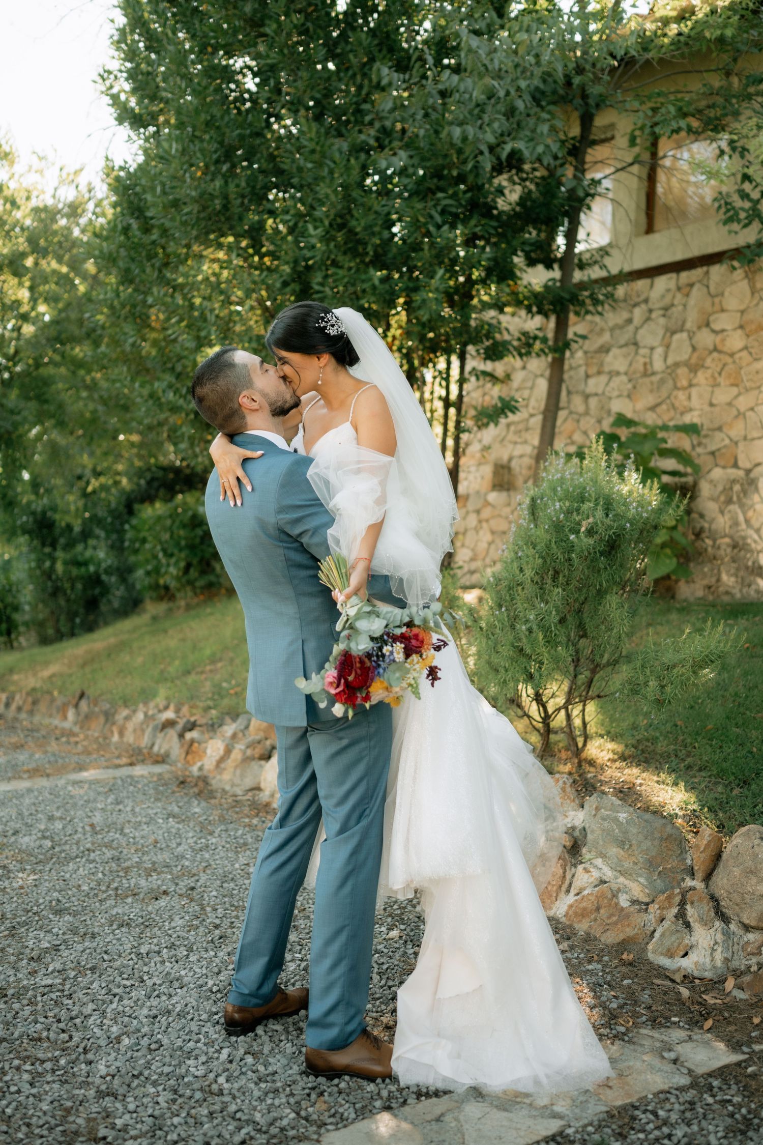 A romantic wedding couple embraces on a gravel path beside a stone wall in a garden setting at sunset.