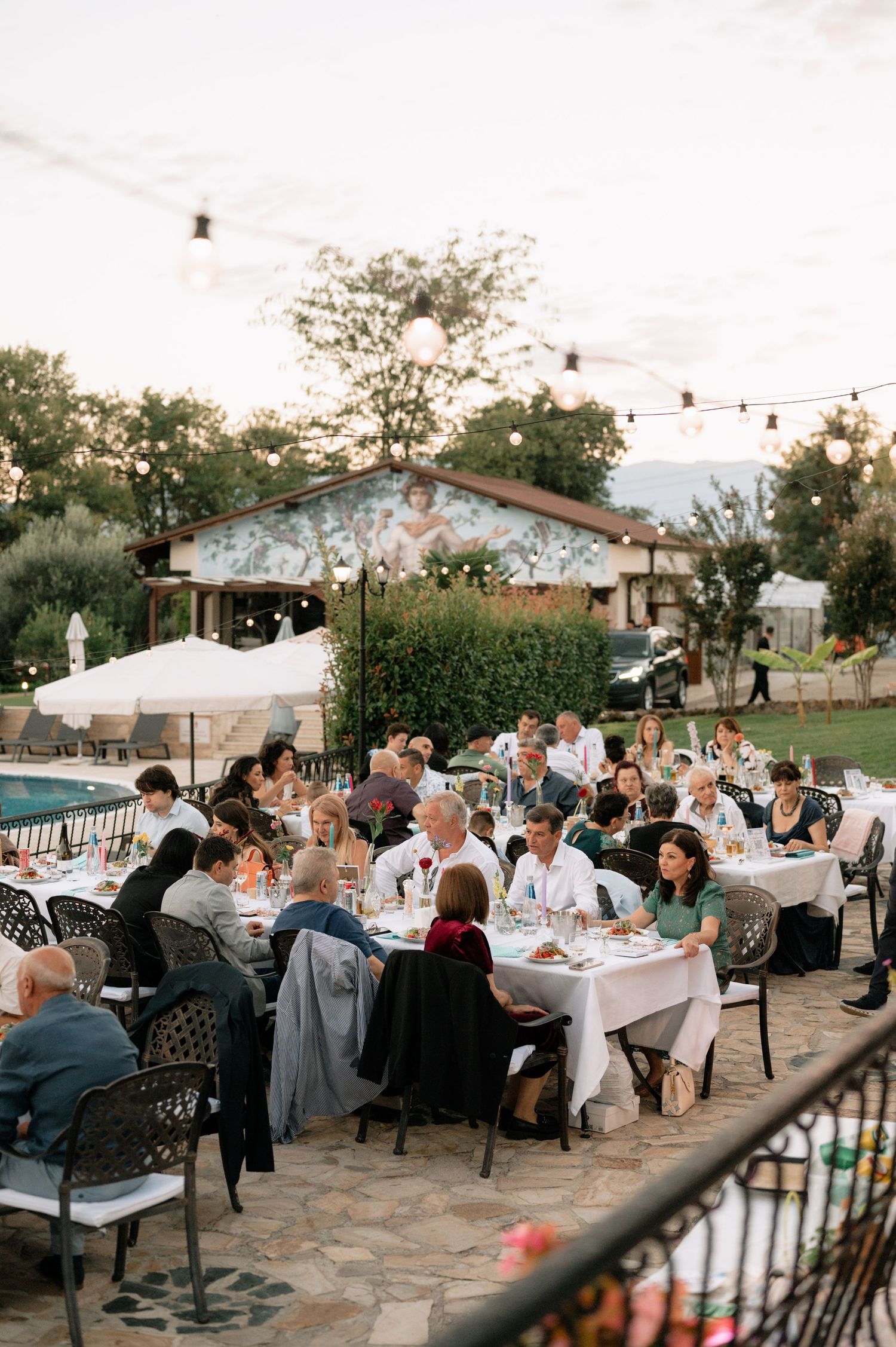Guests seated at elegant outdoor tables during a twilight garden reception with string lights overhead.