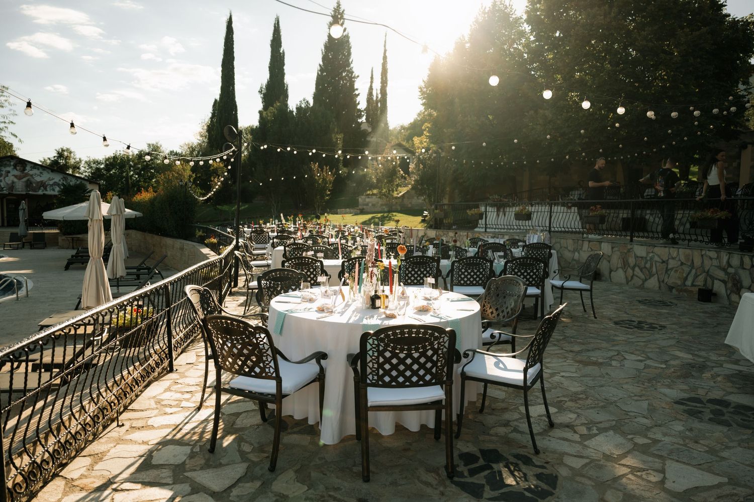 Outdoor wedding reception setup with round tables and chairs on a stone patio at sunset with cypress trees in background.