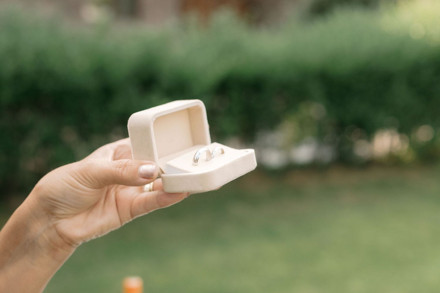 A hand holds a white ceramic teacup in a garden setting with lush green foliage in the background.
