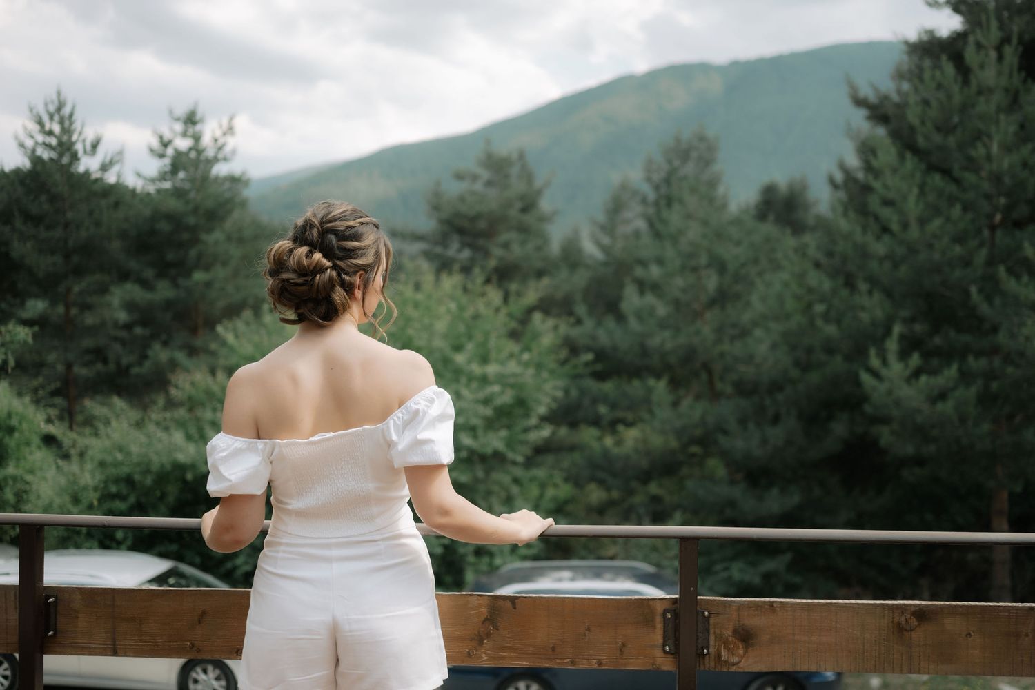 A bride in a white dress stands at a wooden railing overlooking mountains and evergreen trees on a cloudy day.