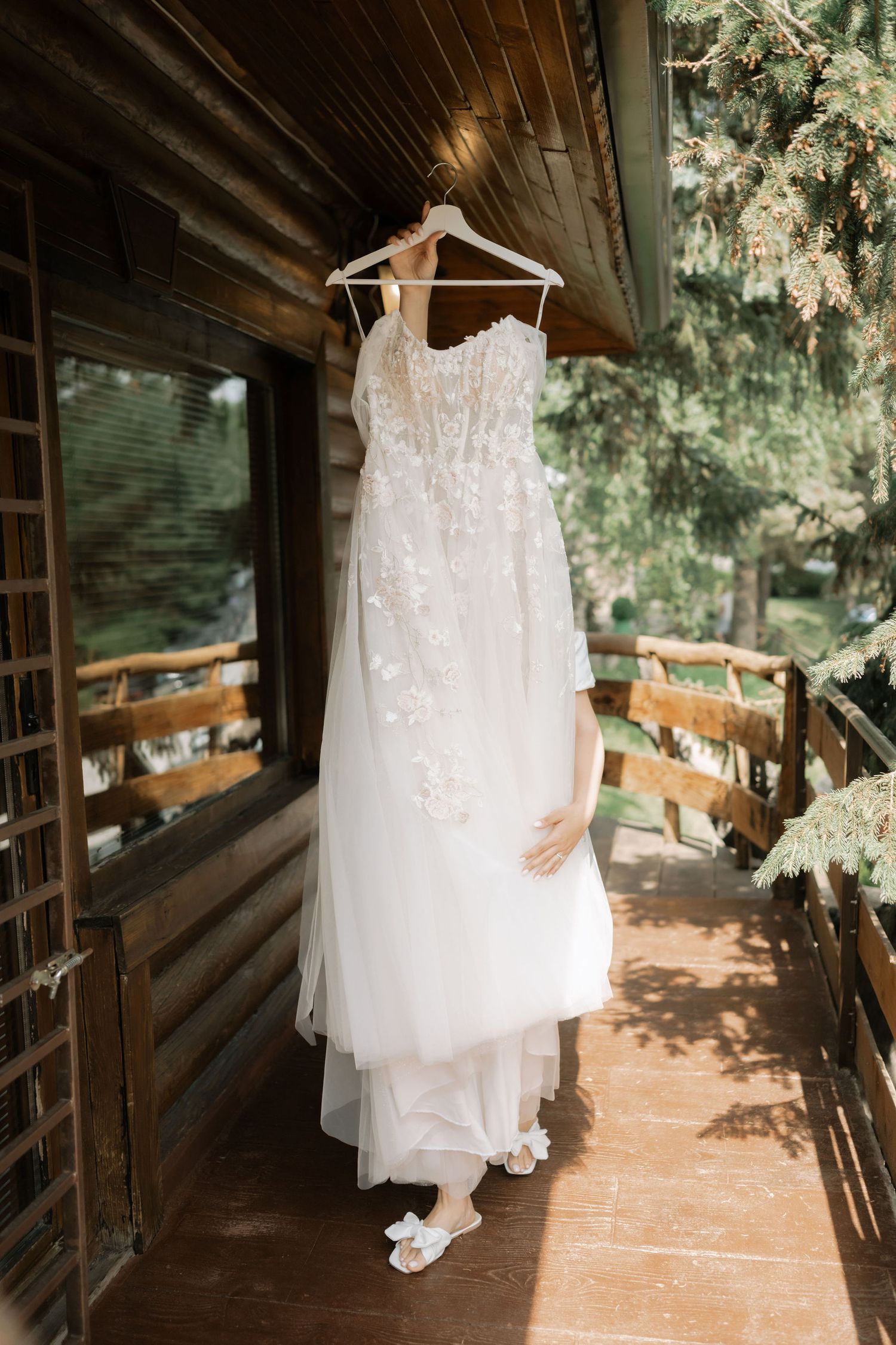 A white lace wedding dress hangs on a wooden hanger against a rustic porch backdrop with soft natural lighting.