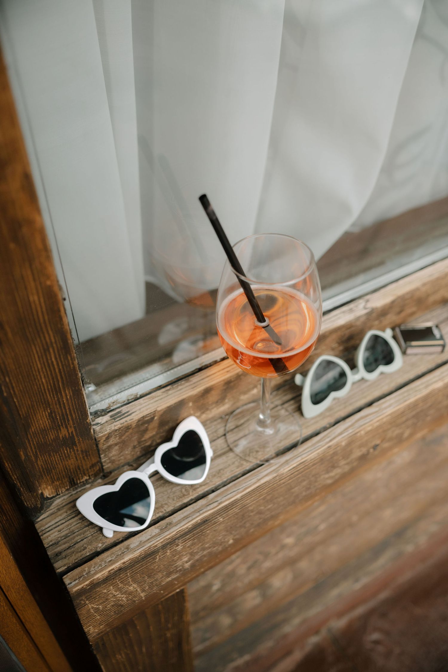 Pink cocktail in wine glass with black straw sits beside white heart-shaped sunglasses on rustic wooden windowsill.