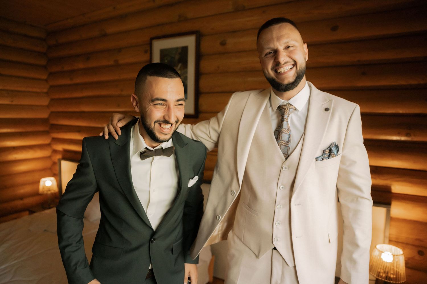 Two people in formal attire smile and embrace in front of a rustic wooden wall with warm lighting.