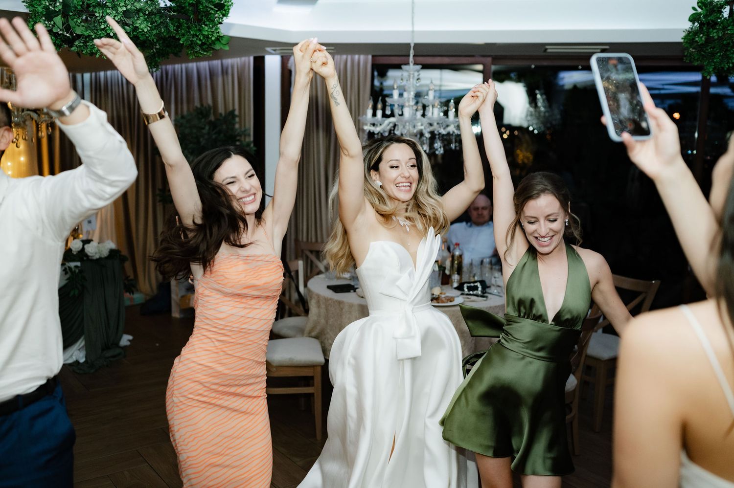 Wedding guests dance and celebrate with raised arms on a dimly lit dance floor during a reception party.