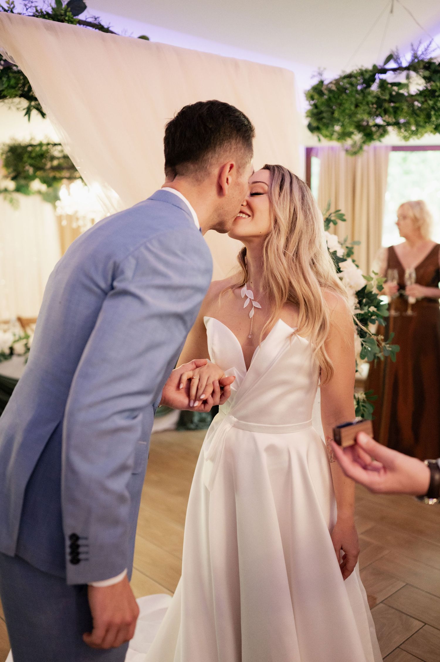 A couple shares their first kiss at an indoor wedding ceremony decorated with greenery and soft lighting.