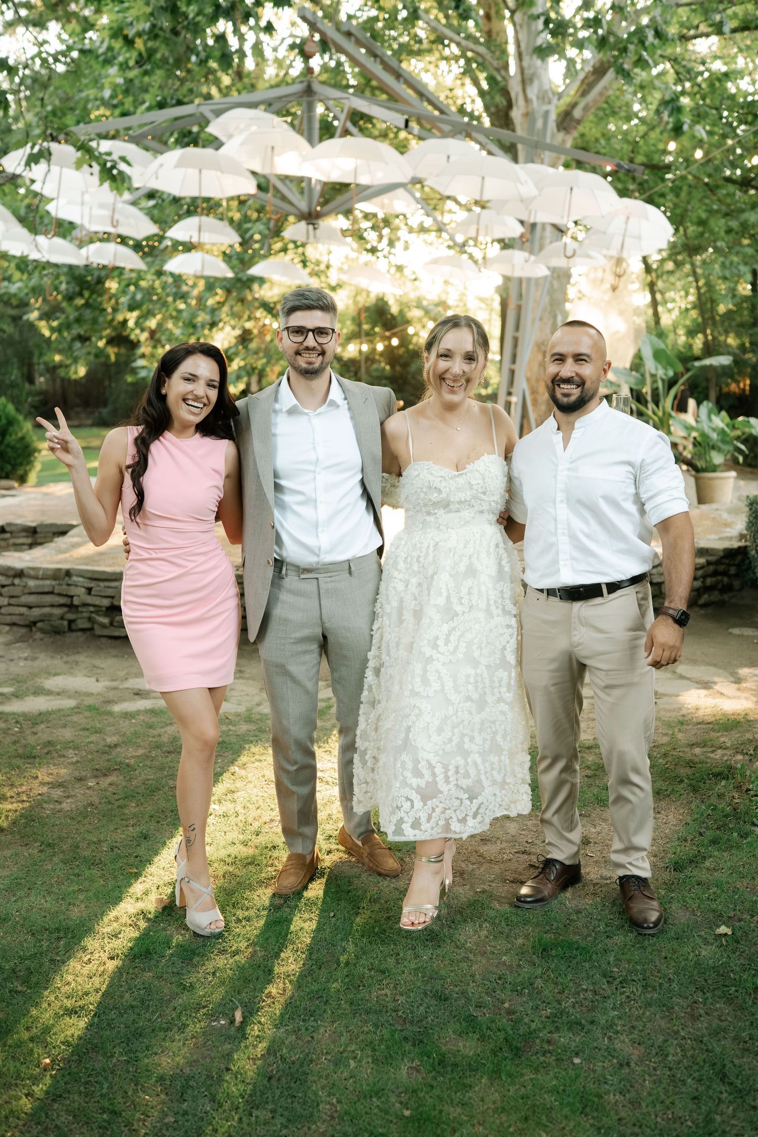Wedding party poses together on grass wearing pink, white, and gray attire in a garden setting.