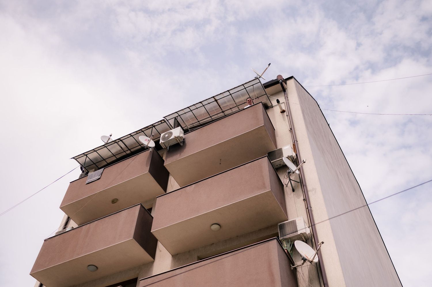 Apartment building exterior with multiple balconies and satellite dishes mounted under cloudy skies.