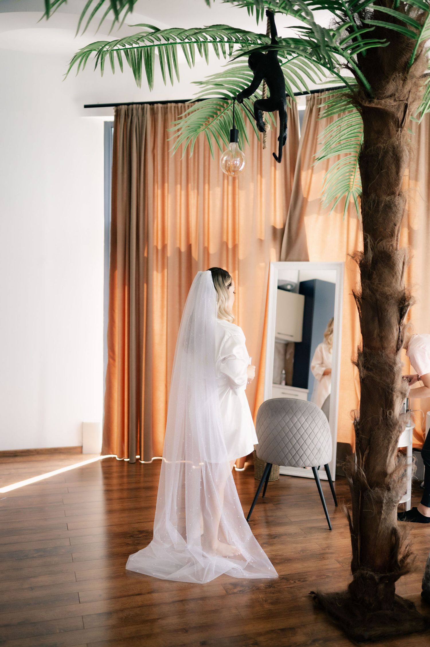 A white sheet draped over a figure creates a ghost-like form in a minimalist room with wooden floors and palm trees.