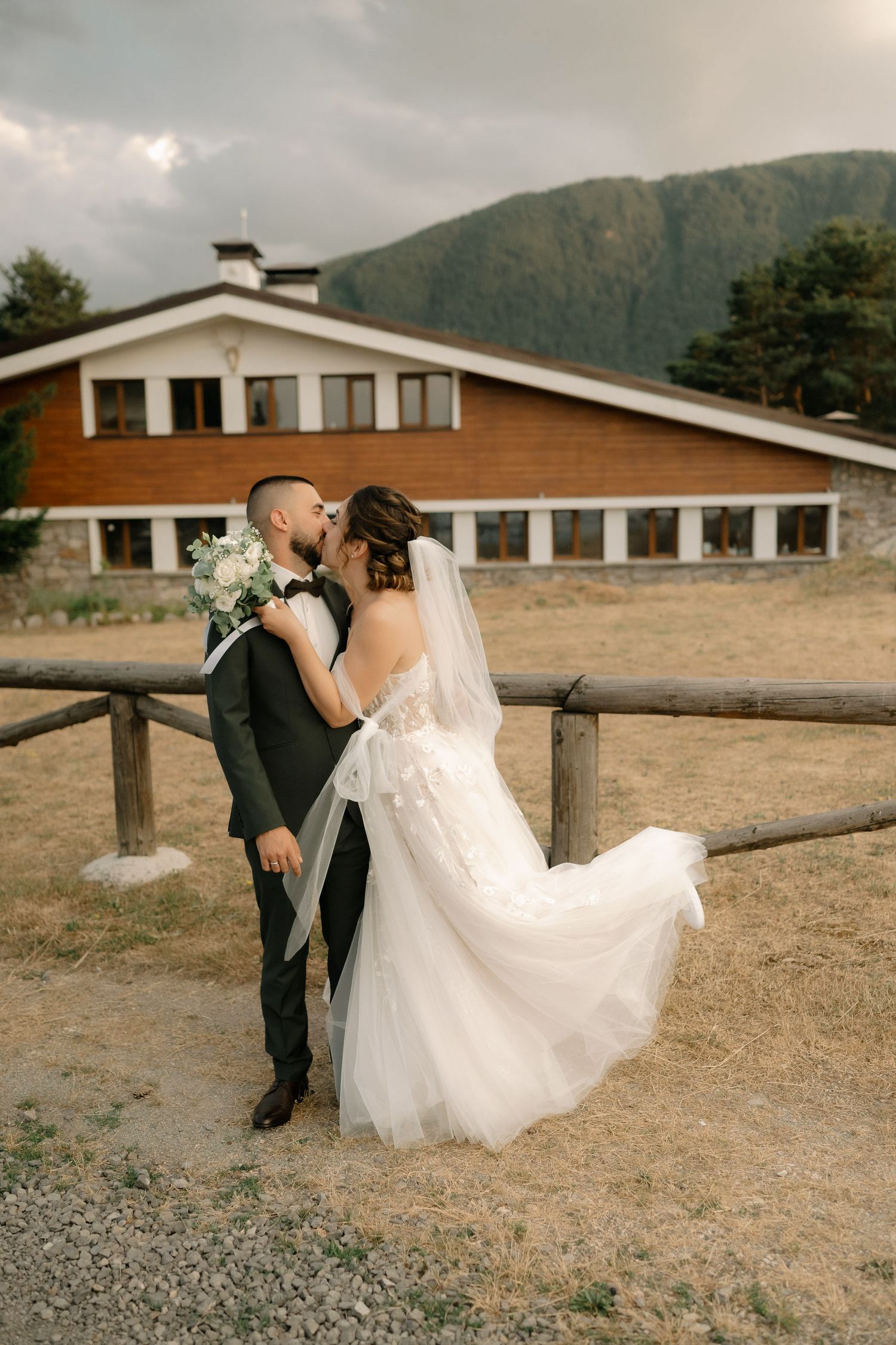 A romantic wedding couple poses along a rustic wooden fence with mountains and a ranch building in the background.