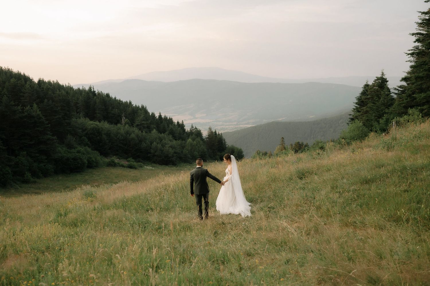 A scenic mountain wedding photoshoot with a couple walking through a grassy meadow during golden hour.
